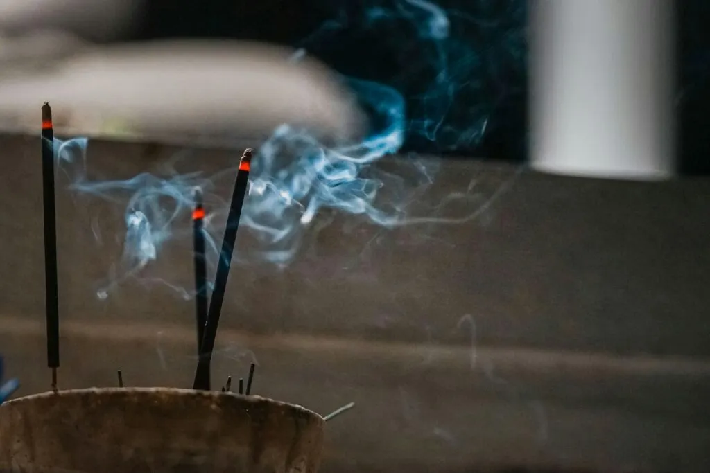 Close-up of burning incense sticks with rising blue smoke indoors, creating a calm atmosphere.