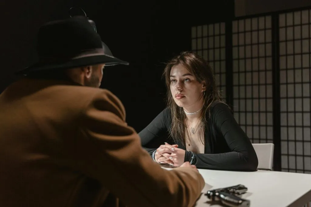 A woman faces a man in a hat during a tense interrogation scene at a table.